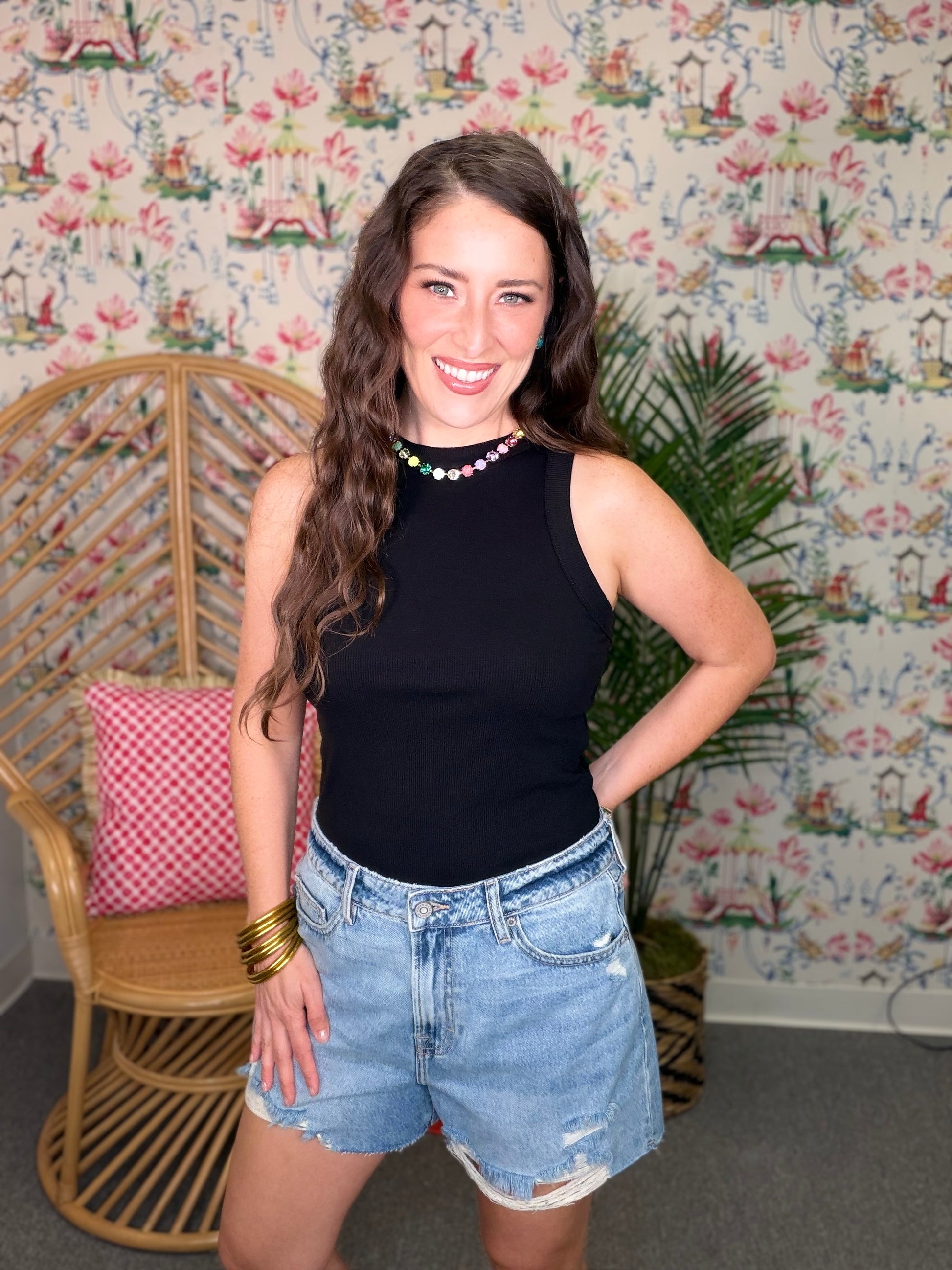 Woman wearing a black sleeveless top and denim shorts standing in a room with floral wallpaper.