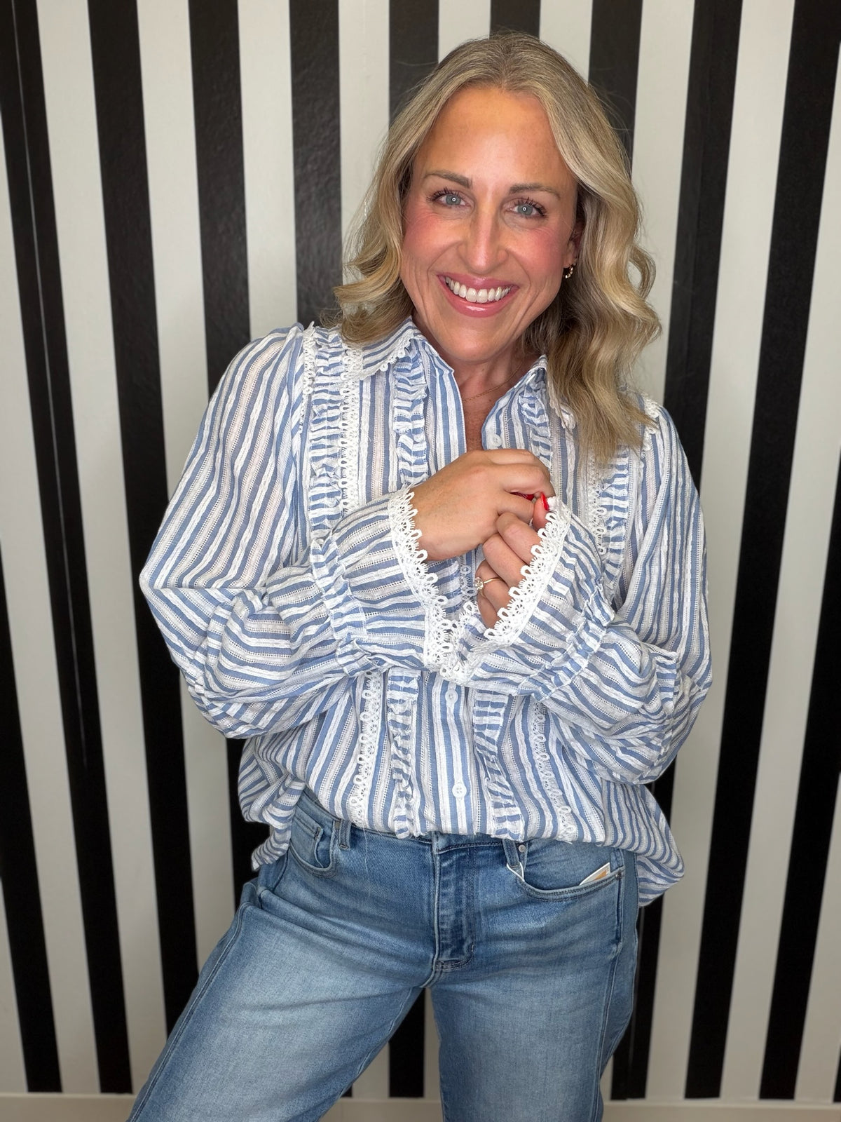 A smiling blonde woman wearing a blue and white vertically striped blouse with white lace trim on the front and cuffs, paired with blue jeans, standing against a black and white striped wall.