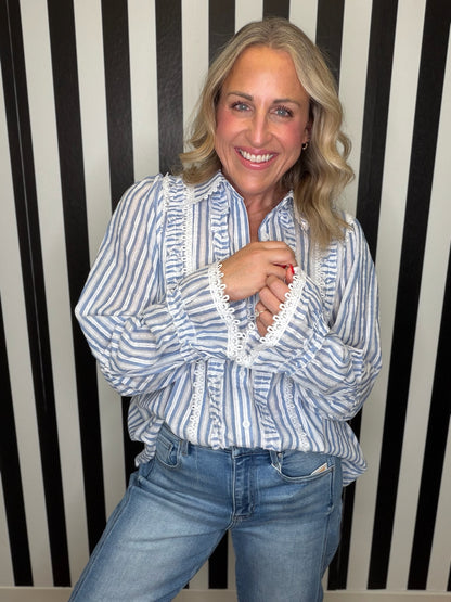 A smiling blonde woman wearing a blue and white vertically striped blouse with white lace trim on the front and cuffs, paired with blue jeans, standing against a black and white striped wall.