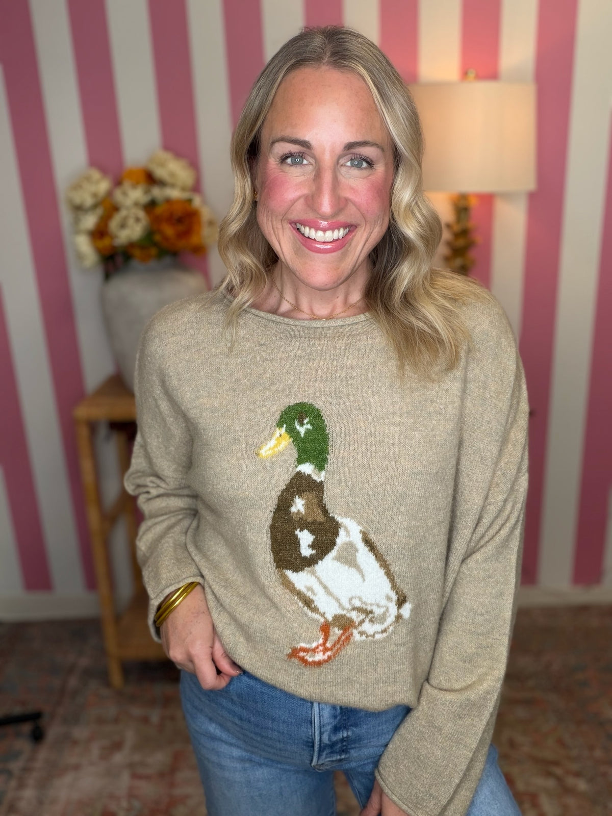 A smiling blonde woman wearing a beige knit sweater with a large embroidered mallard duck design on the front, paired with blue jeans, standing in front of pink and white striped wallpaper.