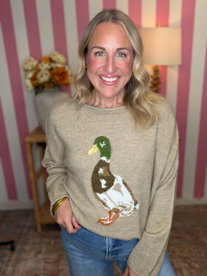A smiling blonde woman wearing a beige knit sweater with a large embroidered mallard duck design on the front, paired with blue jeans, standing in front of pink and white striped wallpaper.