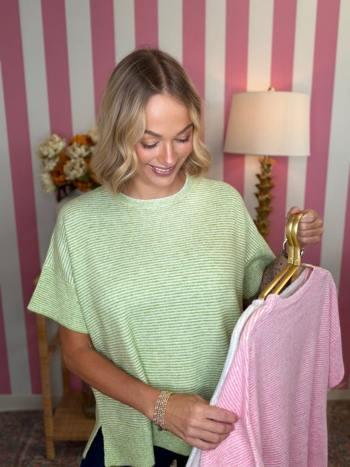 A woman with blonde hair stands in a room with pink and white striped wallpaper, holding up two striped t-shirts on hangers. The shirt on the left is light green, and the shirt on the right is light pink.