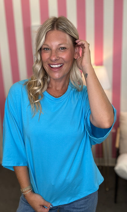 A smiling blonde woman wearing a bright blue short-sleeve t-shirt and blue jeans, standing in front of a pink and white striped wall.