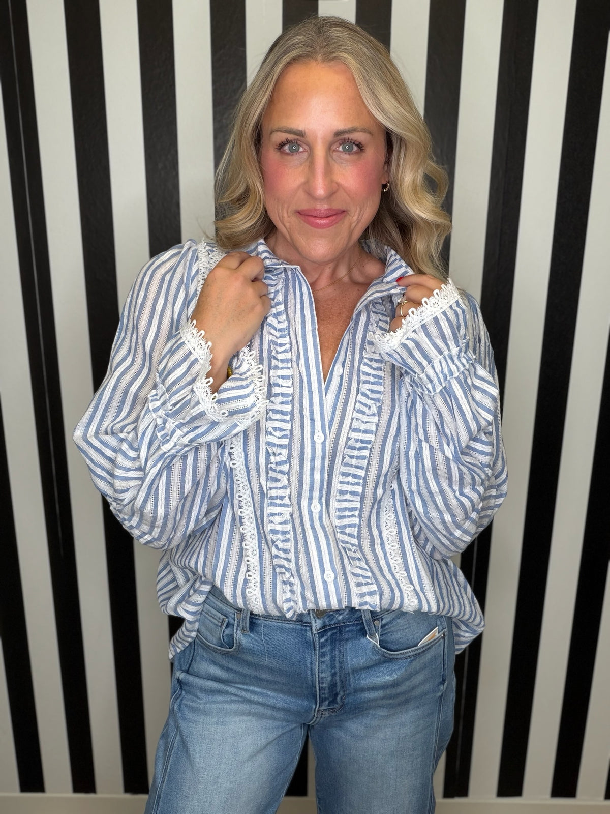 A woman wearing a blue and white vertically striped blouse with white lace trim on the cuffs and front ruffles, paired with blue jeans, standing in front of a black and white striped wall.