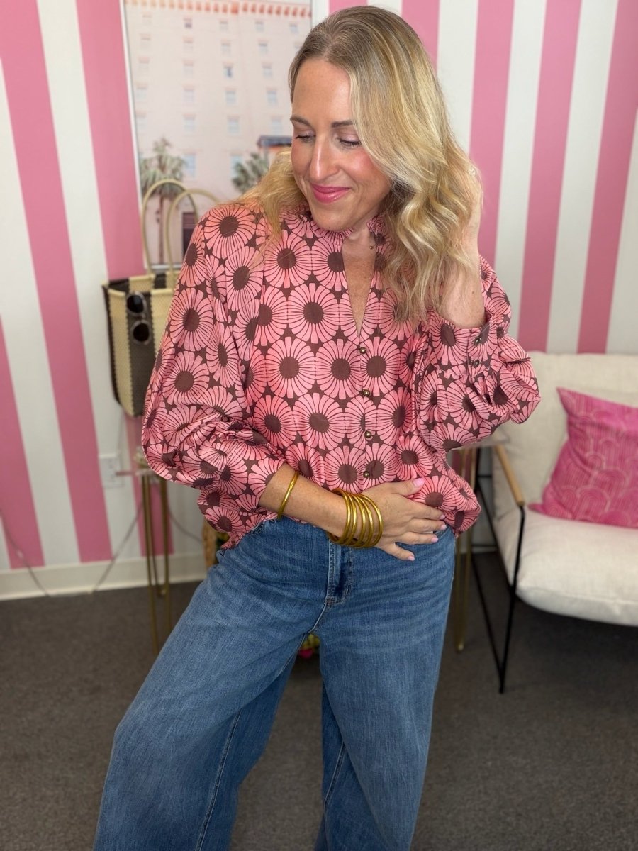 A woman wearing a pink and brown sunburst patterned blouse with blue jeans, standing in front of a pink and white striped wall.