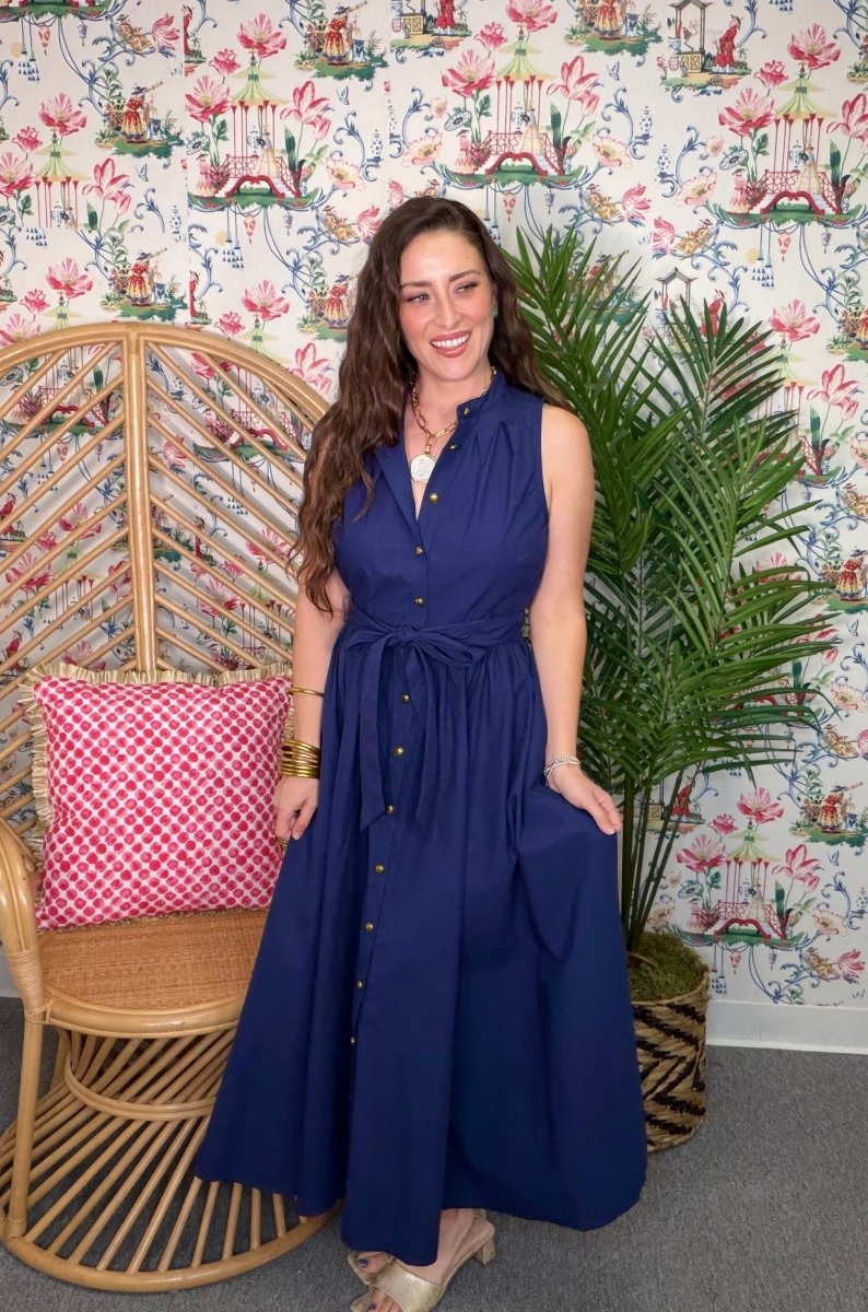 Woman in a blue dress standing in a room with floral wallpaper, wicker chair, and plant.