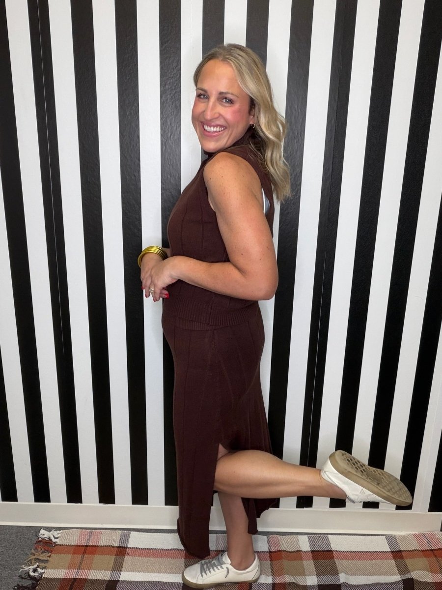 A woman posing in a sleeveless, high-neck, dark brown midi dress with a side slit, paired with white sneakers, standing against a black and white striped wall.