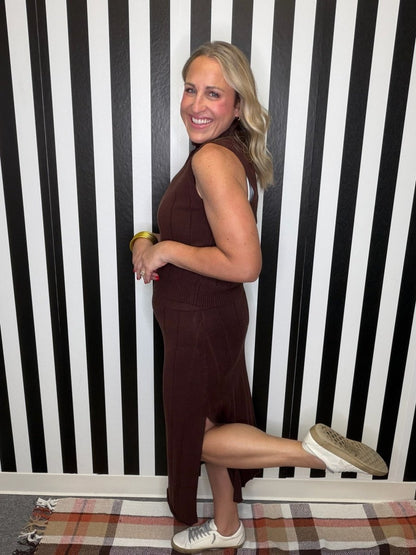 A woman posing in a sleeveless, high-neck, dark brown midi dress with a side slit, paired with white sneakers, standing against a black and white striped wall.