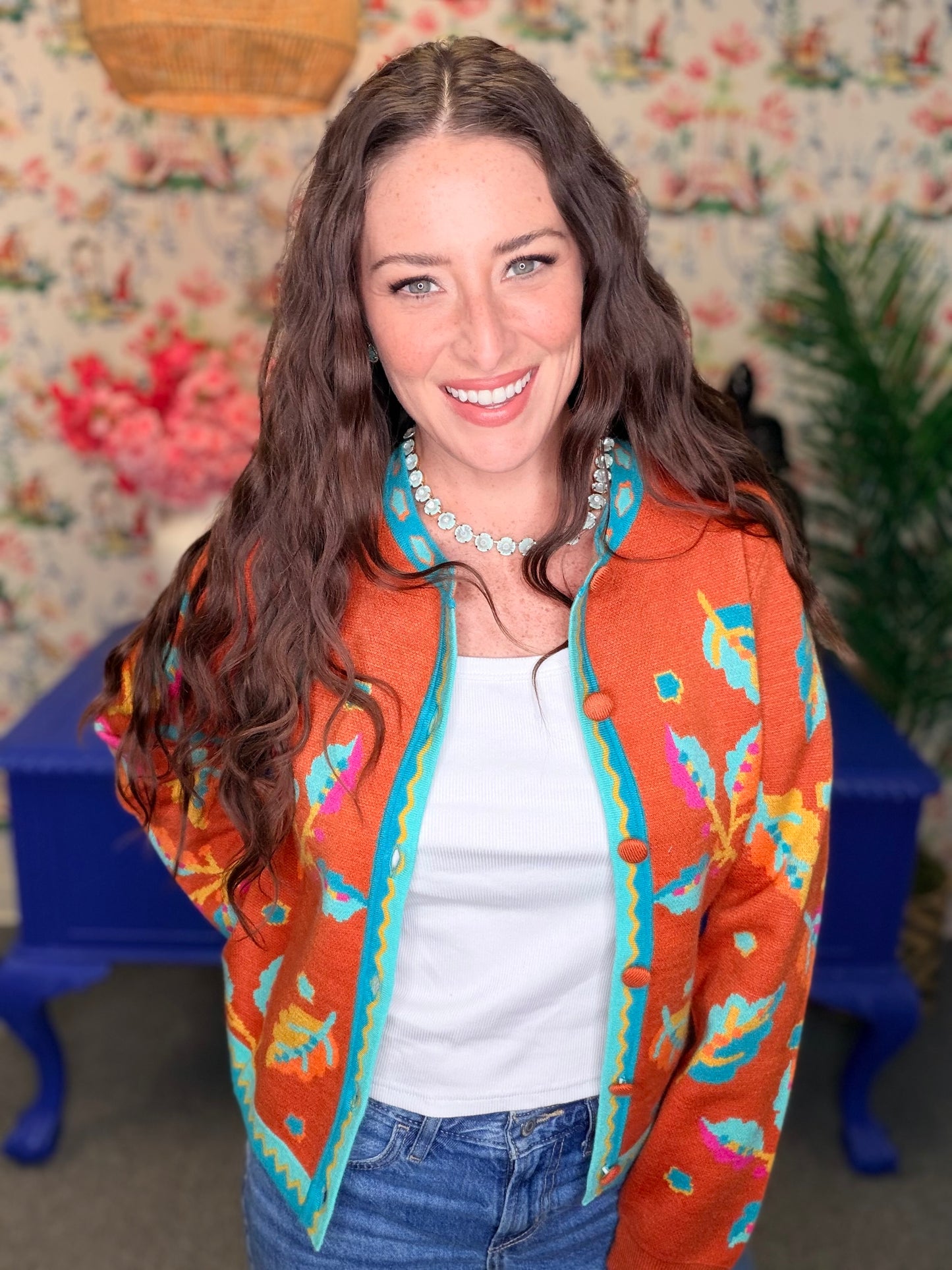 A woman with long brown hair smiling, wearing a vibrant orange knit cardigan with colorful floral patterns and turquoise trim, styled with a white top and blue jeans.