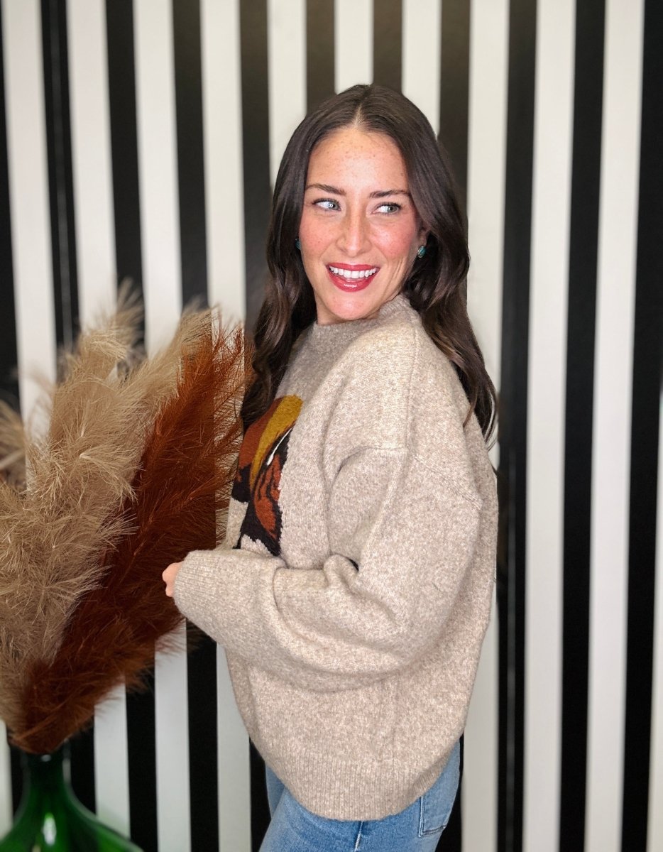 A woman wearing a beige knit crewneck sweater with a colorful graphic on the front, standing in front of a black and white striped wall and holding a decorative pampas grass bundle.