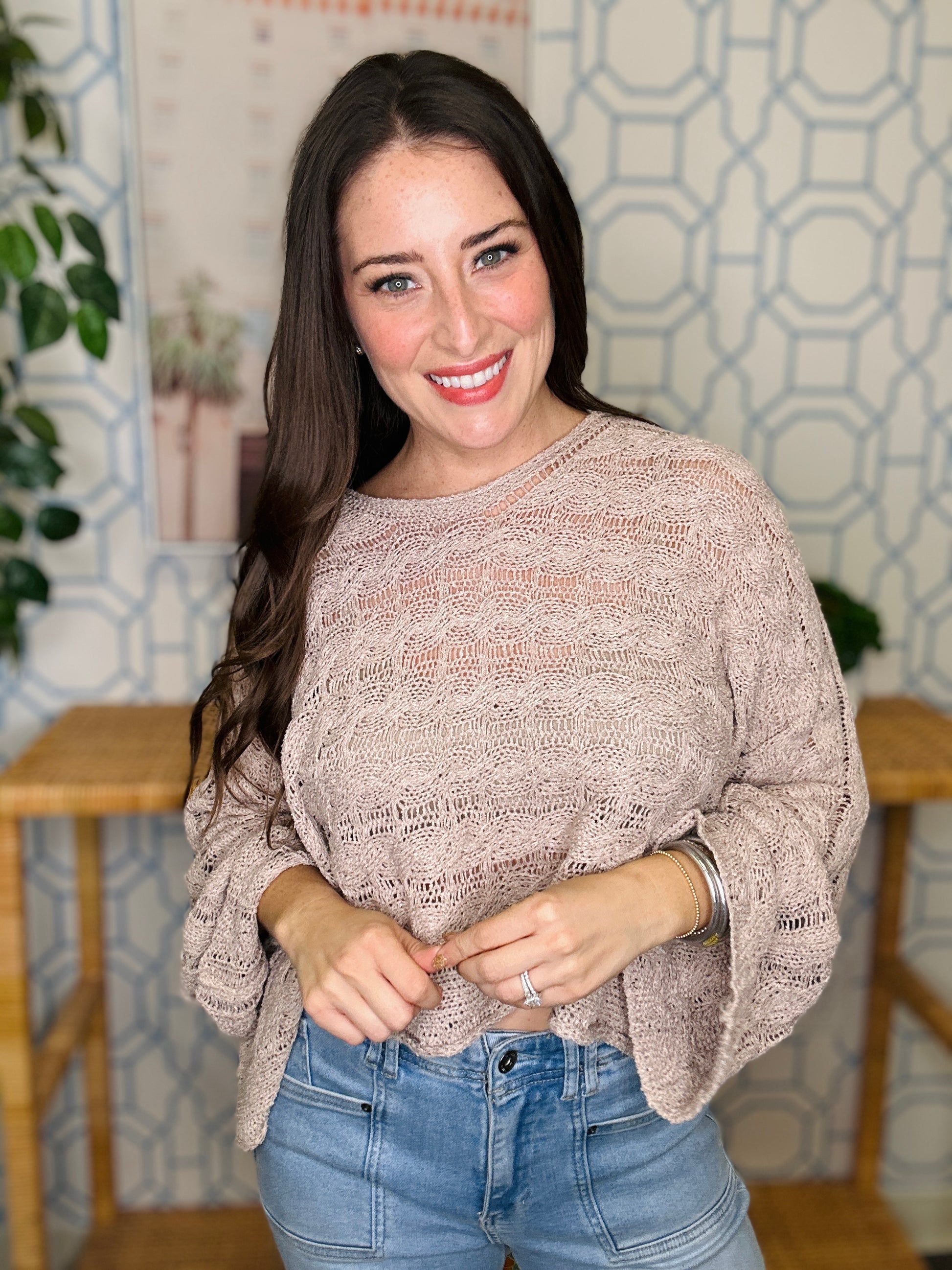Woman wearing a beige sweater and blue jeans standing indoors with a decorative wall in the background.