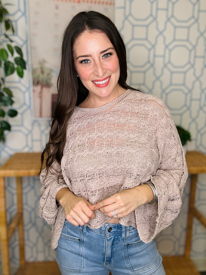 Woman wearing a beige sweater and blue jeans standing indoors with a decorative wall in the background.