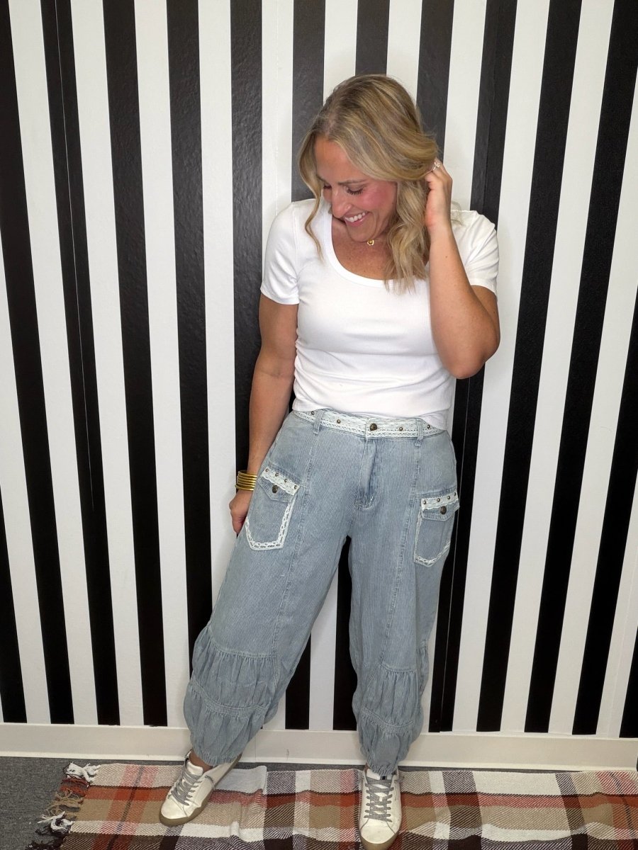 A woman wearing a white t-shirt and light wash denim baggy pants with ruffle details, standing in front of a black and white striped wall.