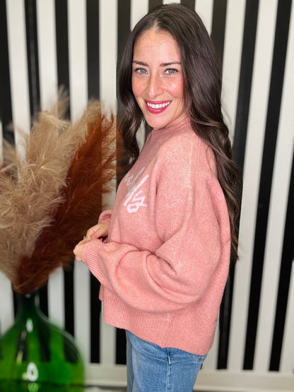A woman with long dark brown hair smiles while wearing a fuzzy dusty pink crewneck sweater with a white graphic on the chest, paired with blue jeans. She is standing in front of a black and white striped background with a green vase and pampas grass visible on the left.