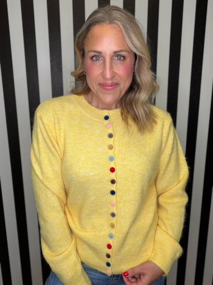 A woman with blonde hair wearing a bright yellow knit sweater with colorful buttons down the front, standing against a black and white vertically striped background.