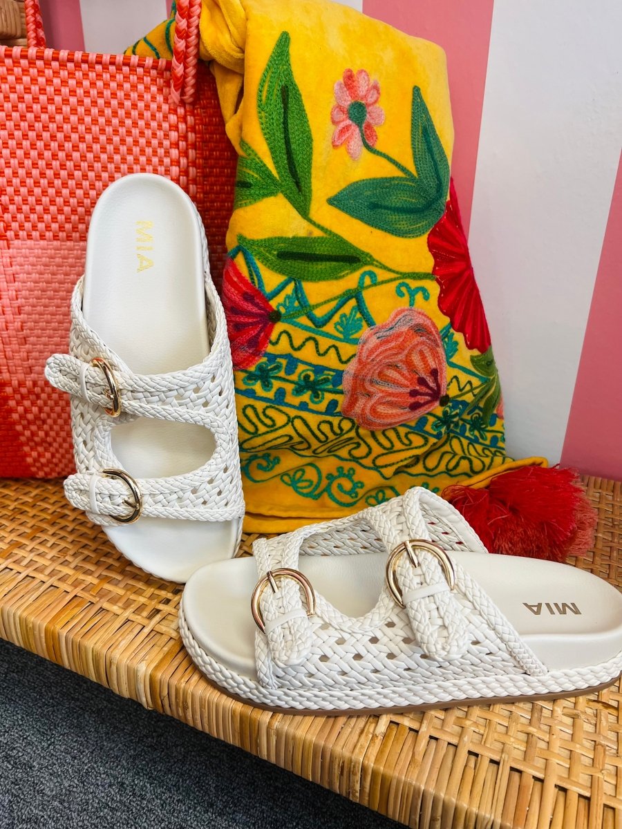 White sandals with gold straps on a woven surface next to a colorful bag.