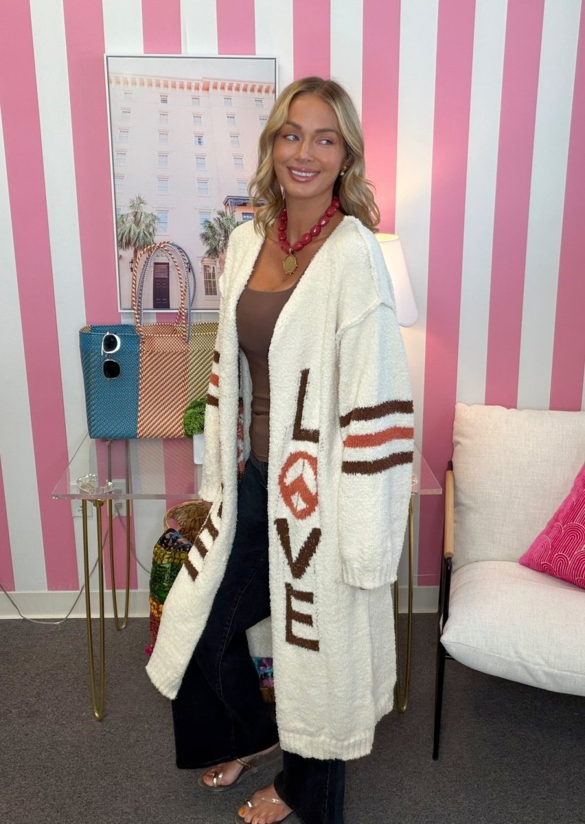 A woman stands in a room with pink and white striped walls wearing a cream bouclé cardigan with brown and orange lettering that reads LOVE. She is also wearing a brown top, dark pants, and a red necklace.