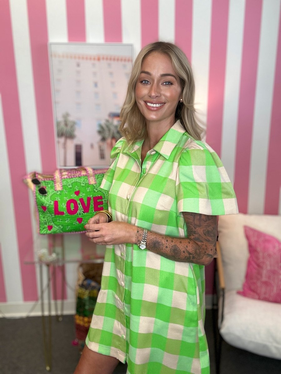 A woman with blonde hair stands in a room with pink and white striped wallpaper, wearing a green and white checkered short-sleeve collared dress and holding a green glittery bag with pink 'LOVE' lettering.