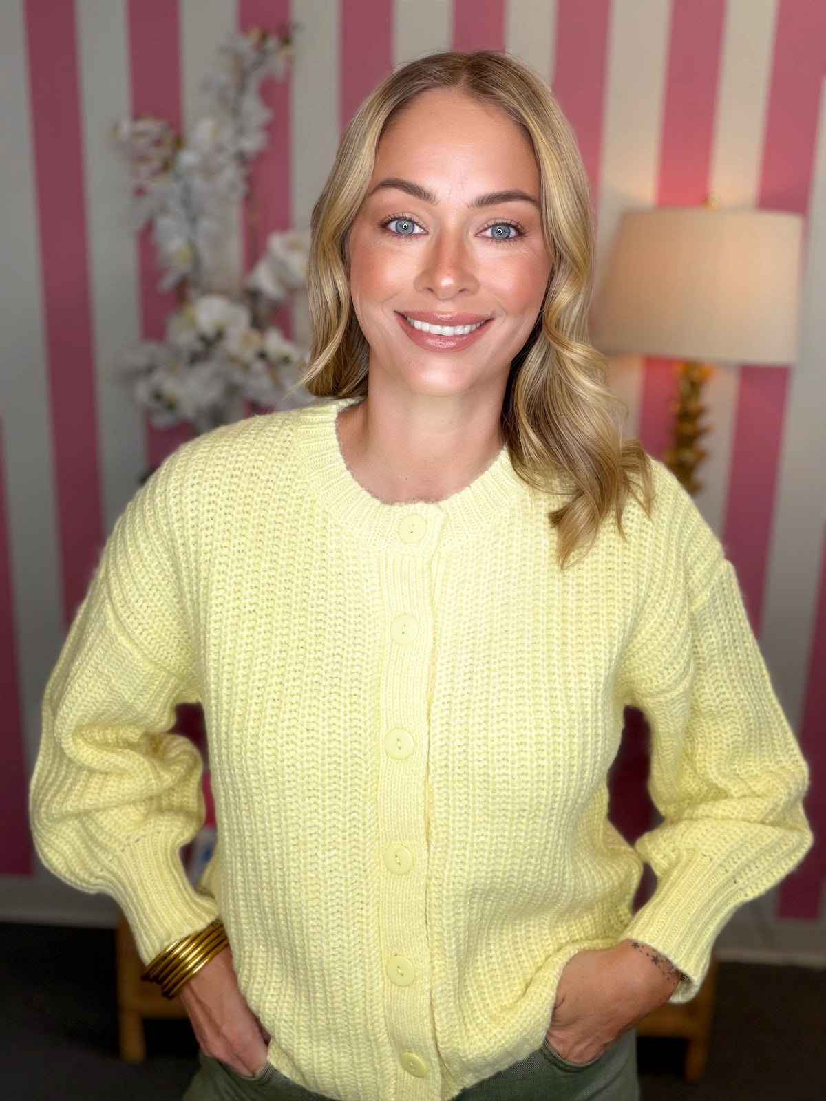 A smiling blonde woman wearing a pale yellow chunky knit button-up cardigan sweater, standing in front of a pink and white striped wall with a lamp in the background.
