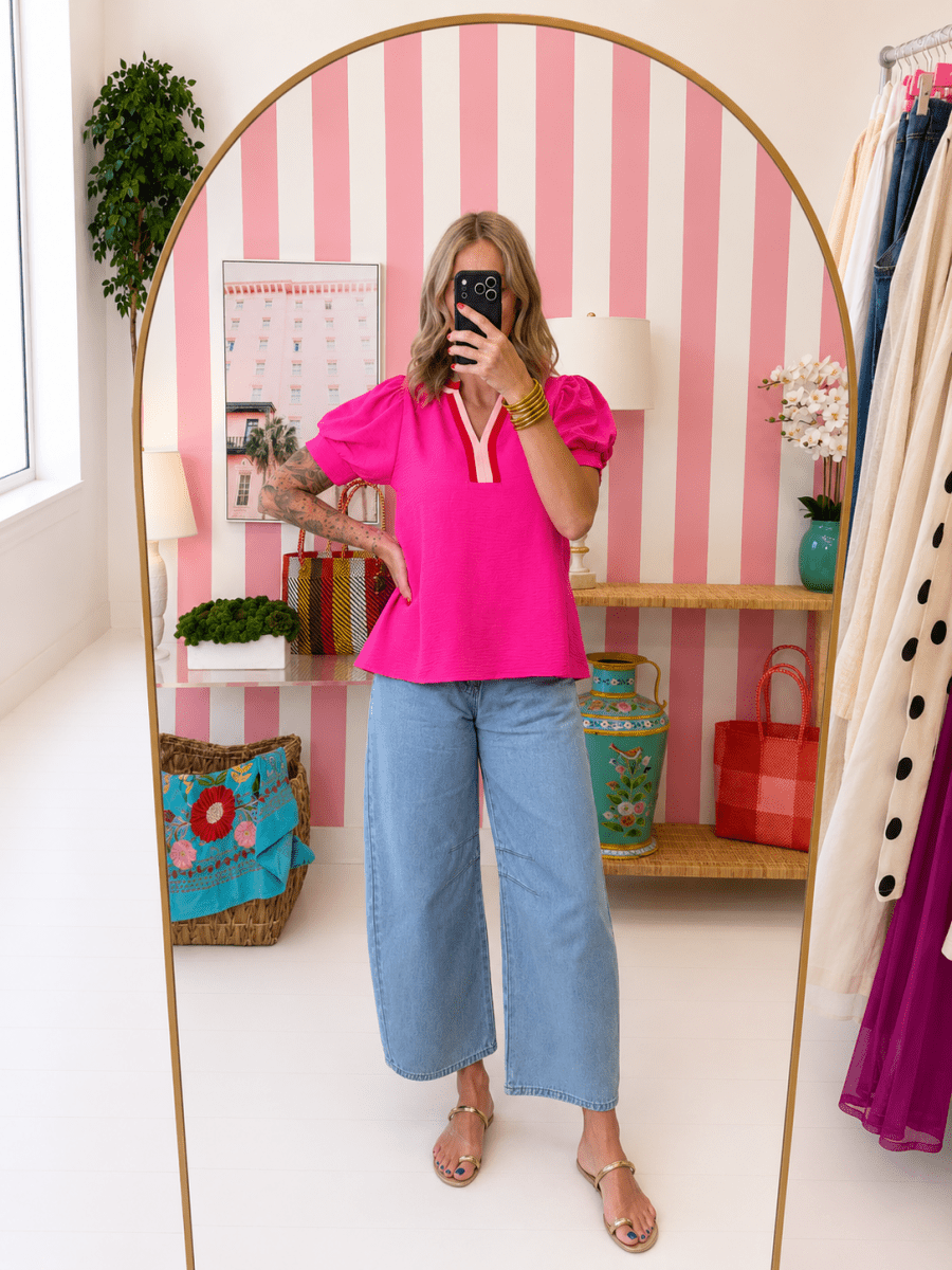 Woman taking a mirror selfie wearing a bright pink top and blue jeans in a room with striped walls and decorative items.