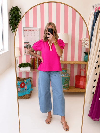 Woman taking a mirror selfie wearing a bright pink top and blue jeans in a room with striped walls and decorative items.