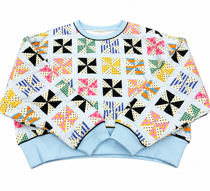 A long-sleeve crewneck sweatshirt featuring a colorful retro patchwork quilt pattern on a white background, with light blue ribbed cuffs and hem.