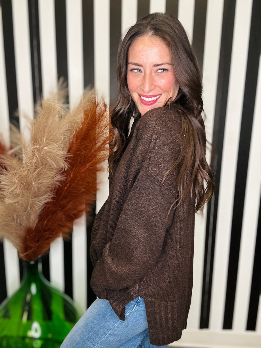 A woman wearing a brown knit sweater and blue jeans, smiling and looking over her shoulder in front of a black and white striped background with a decorative pampas grass arrangement.
