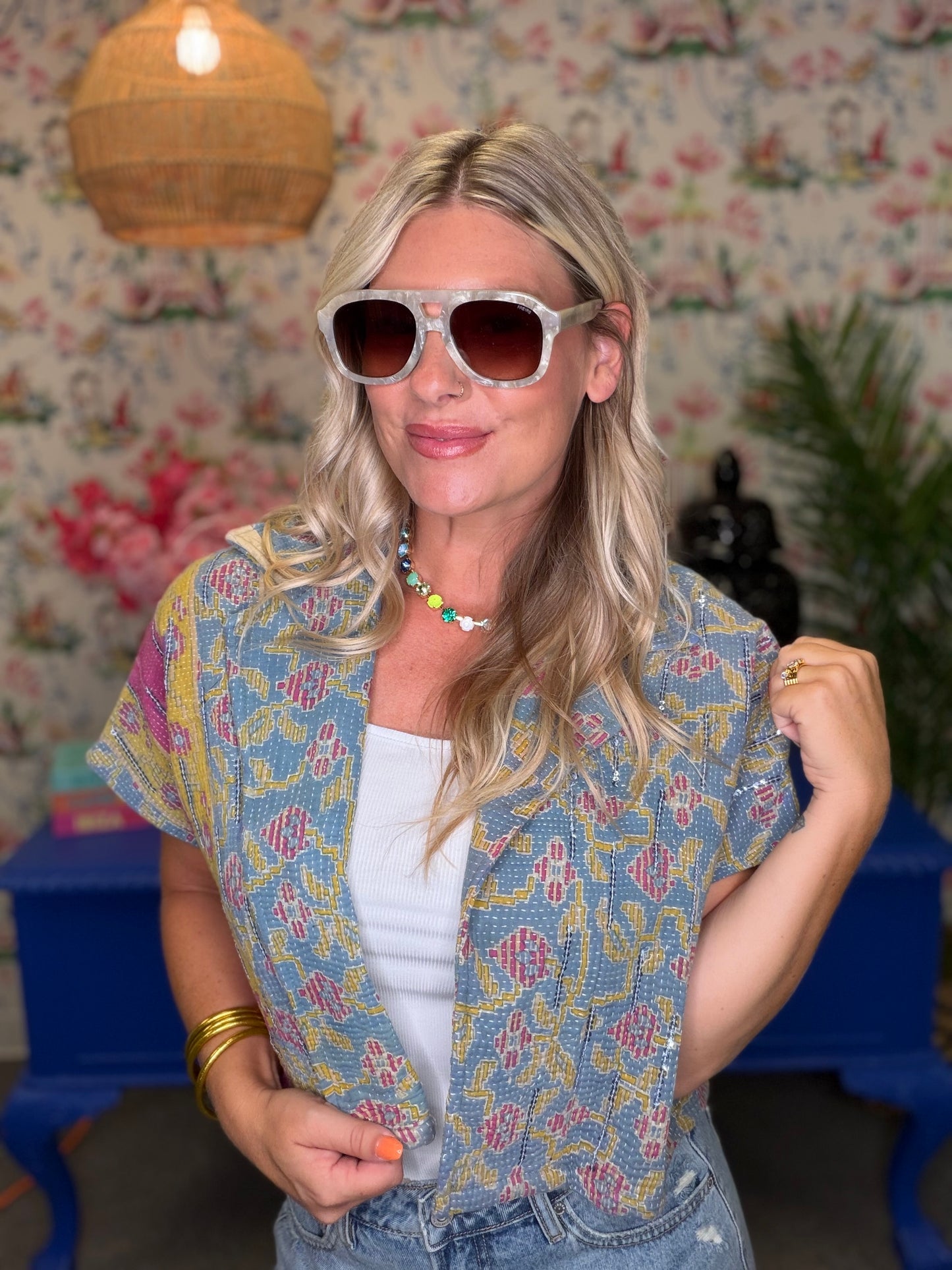 A woman wearing a light blue short-sleeve button-up shirt with a yellow and pink bohemian pattern, styled over a white tank top and denim shorts. She is wearing white-framed sunglasses and standing in front of a floral wallpaper background with a hanging lamp.