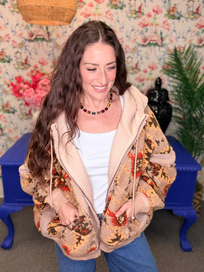 A woman wearing a beige sherpa-lined zip-up hoodie jacket with a brown, red, and black floral pattern on the exterior, styled with a white tank top and blue jeans in a vintage-style room with floral wallpaper.