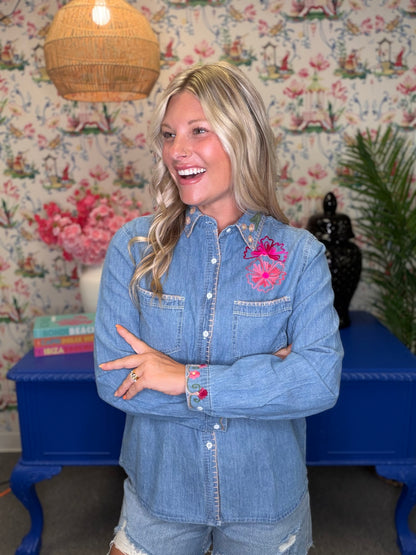A smiling blonde woman wearing a light blue denim button-up shirt with pink floral embroidery on the chest, standing in front of a floral wallpaper background with a blue side table.