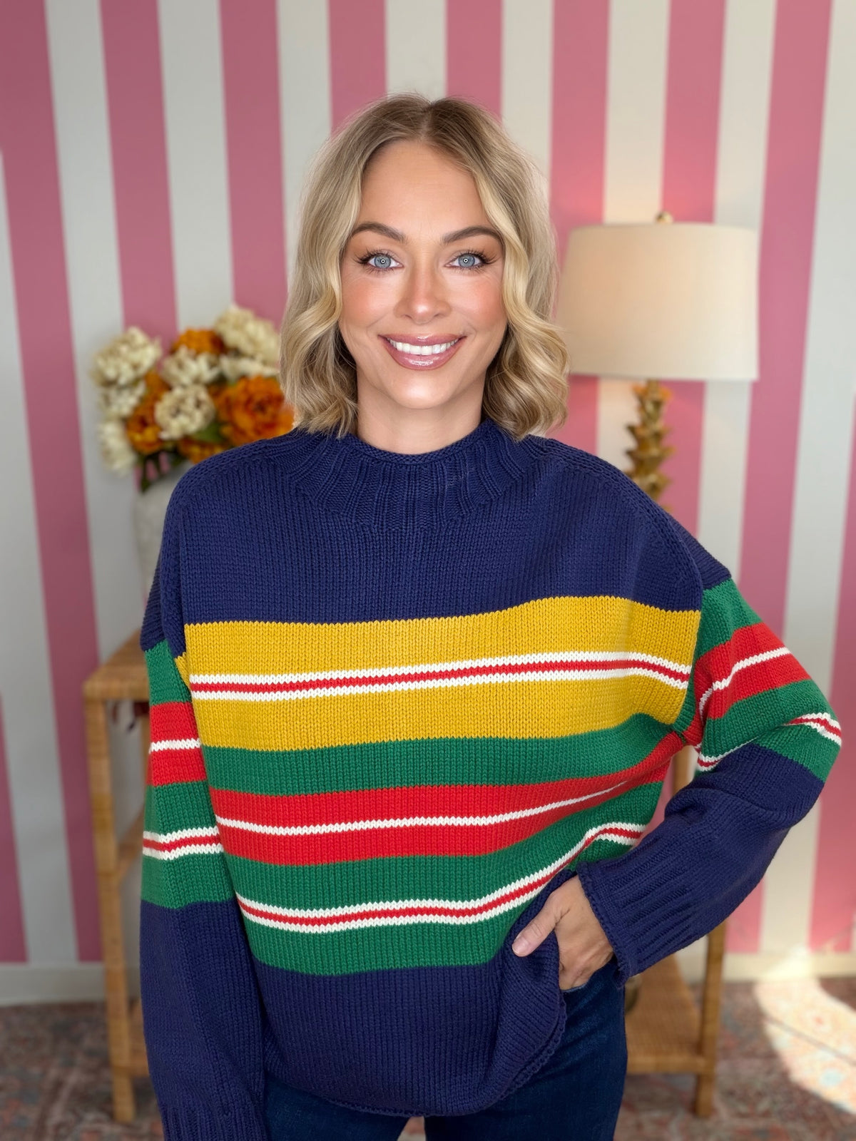 A smiling woman wearing a navy blue knit sweater with bold horizontal stripes in yellow, green, red, and white, posed in front of a pink and white striped wall.
