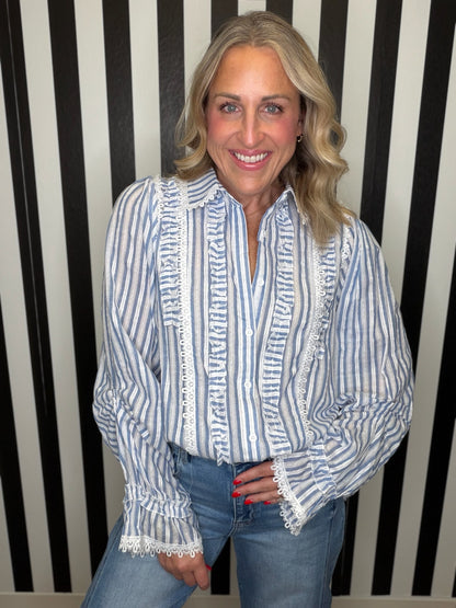 A smiling blonde woman wearing a blue and white vertically striped blouse with white lace trim and blue jeans, standing in front of a black and white striped wall.