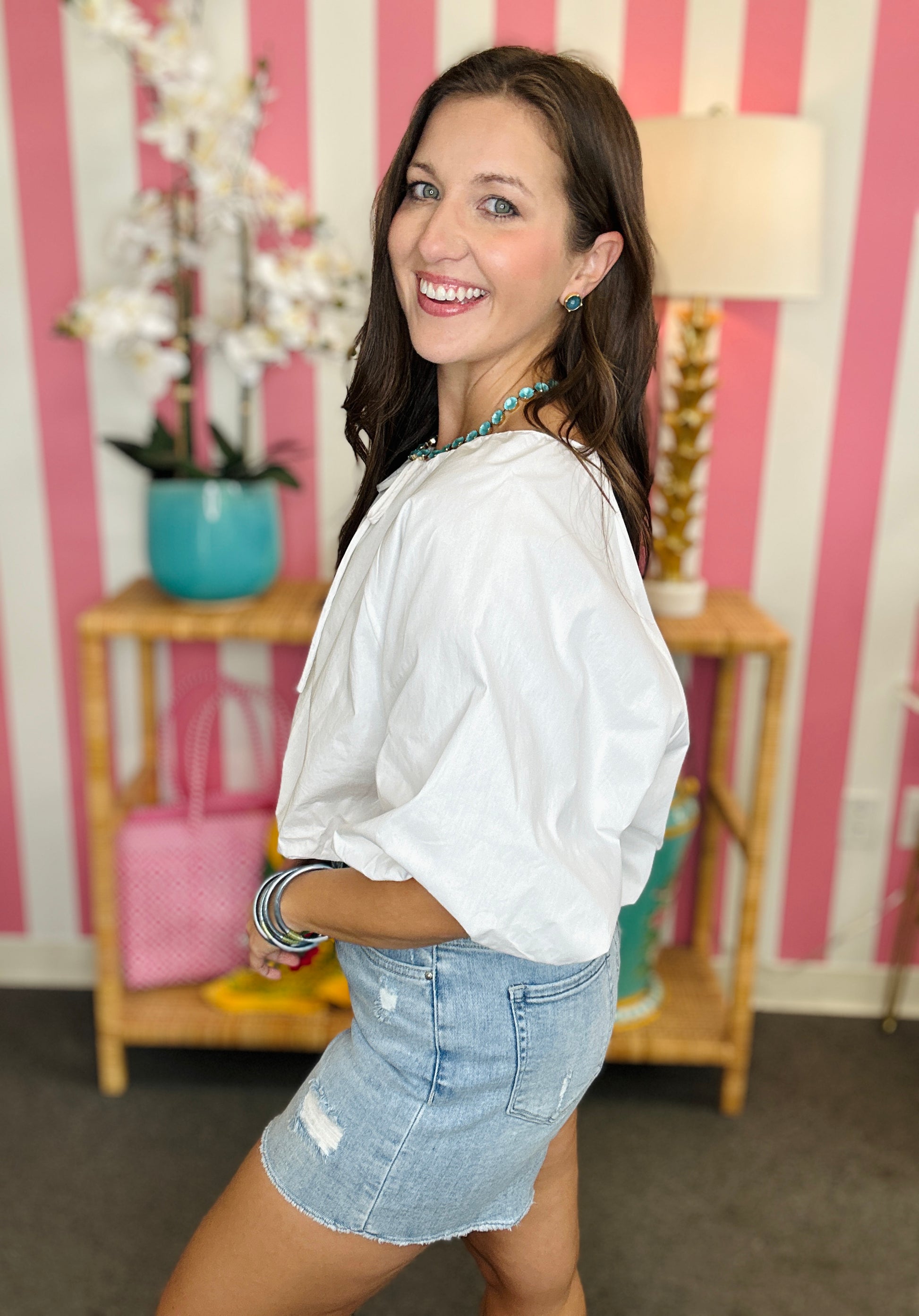Woman in a white blouse and denim shorts standing in a room with striped walls and a lamp.