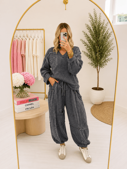 A woman takes a mirror selfie in a boutique wearing a matching charcoal gray lounge set consisting of a long-sleeve V-neck top and wide-leg jogger pants. The boutique features a gold-framed arch mirror, a clothing rack with pink and cream shirts, and a potted plant in the background.