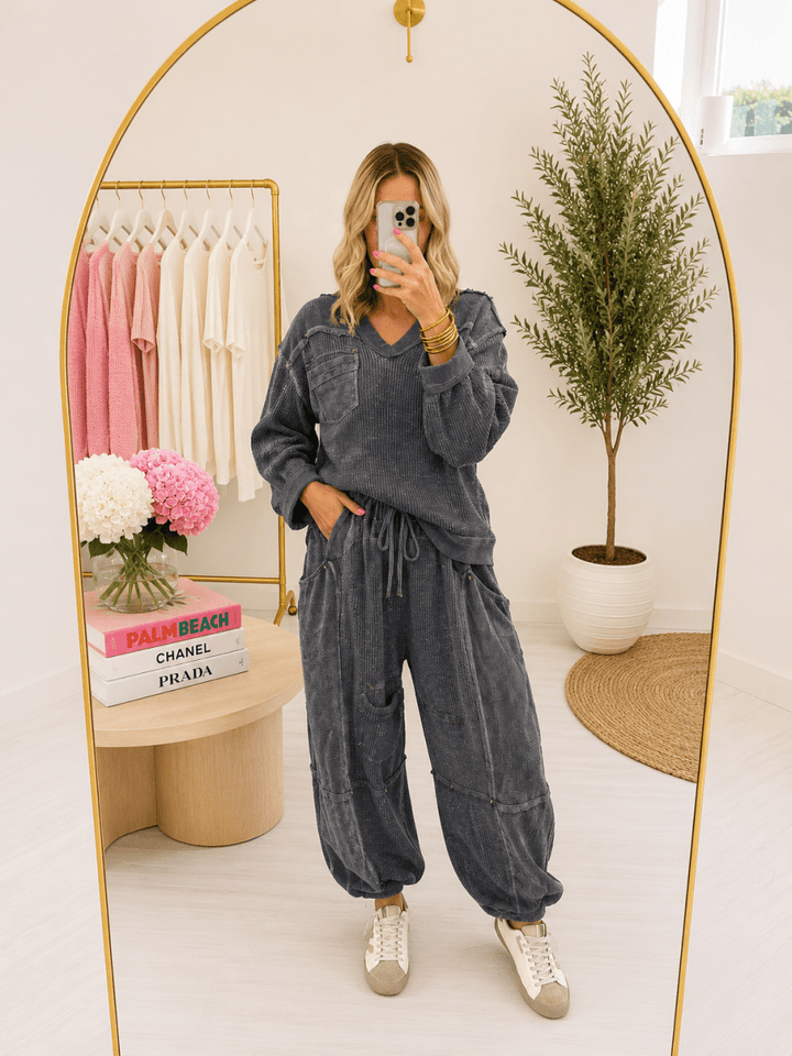 A woman takes a mirror selfie in a boutique wearing a matching charcoal gray lounge set consisting of a long-sleeve V-neck top and wide-leg jogger pants. The boutique features a gold-framed arch mirror, a clothing rack with pink and cream shirts, and a potted plant in the background.