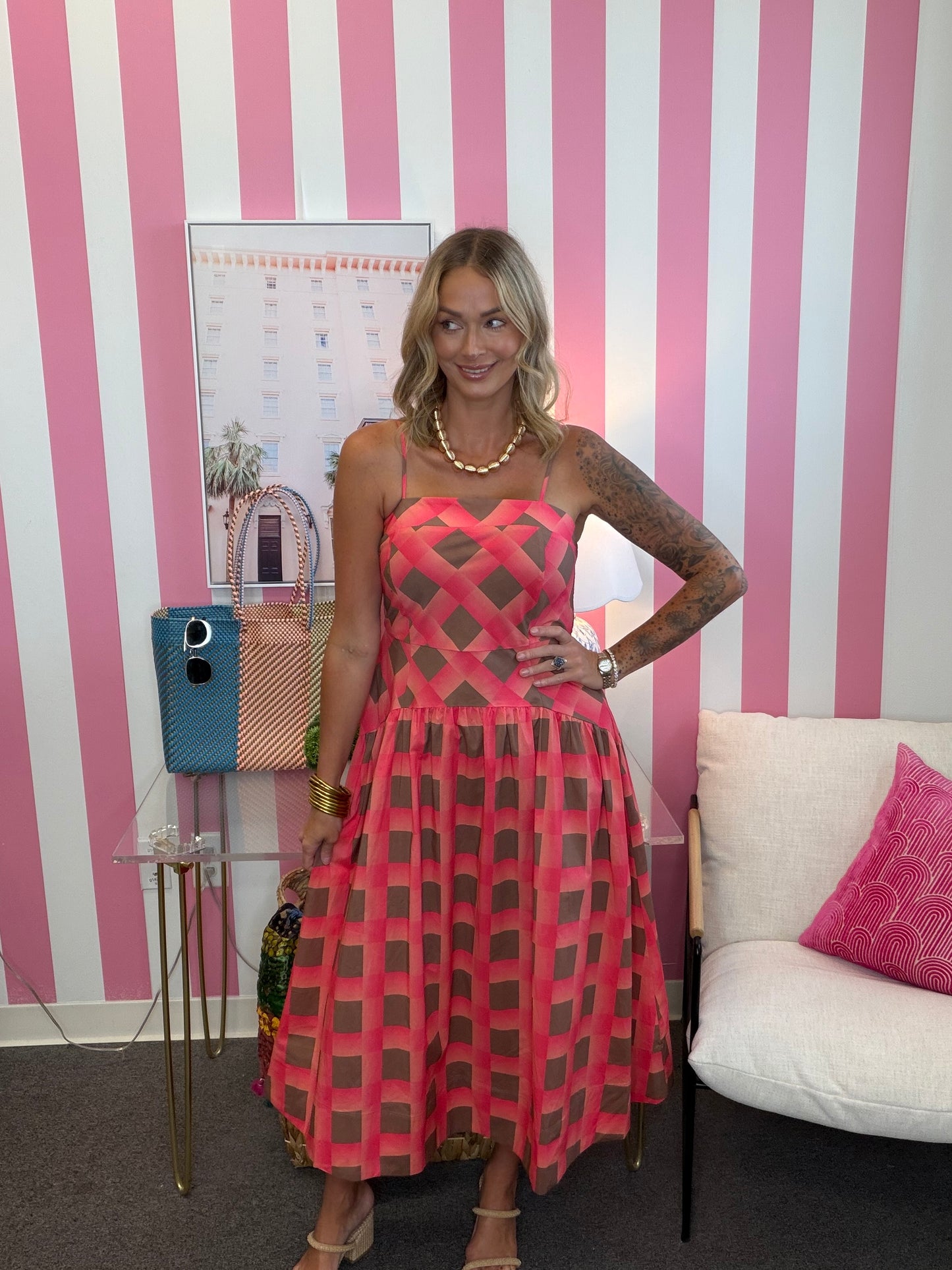 A woman models a pink and brown geometric patterned midi dress with sheer mesh panels, standing in front of a pink and white striped wall.