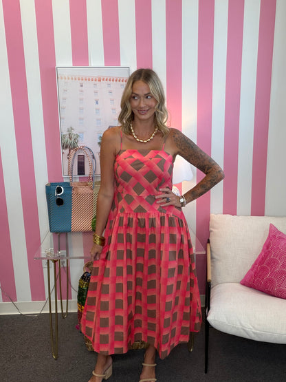 A woman models a pink and brown geometric patterned midi dress with sheer mesh panels, standing in front of a pink and white striped wall.
