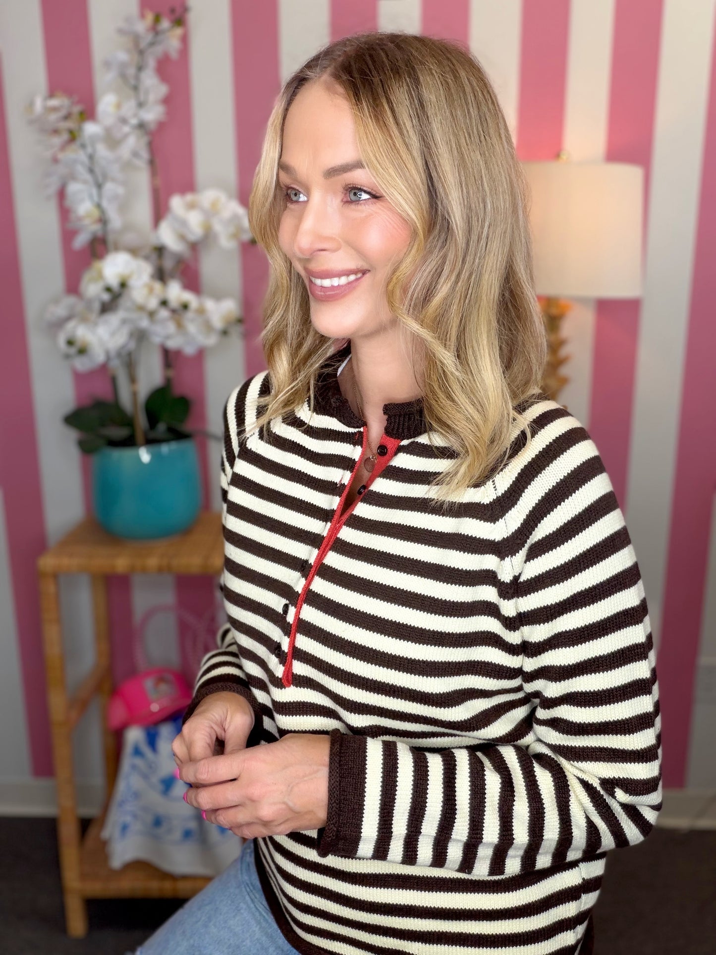 A woman with blonde hair wearing a brown and cream striped quarter-zip sweater, sitting in front of a pink and white striped wall.