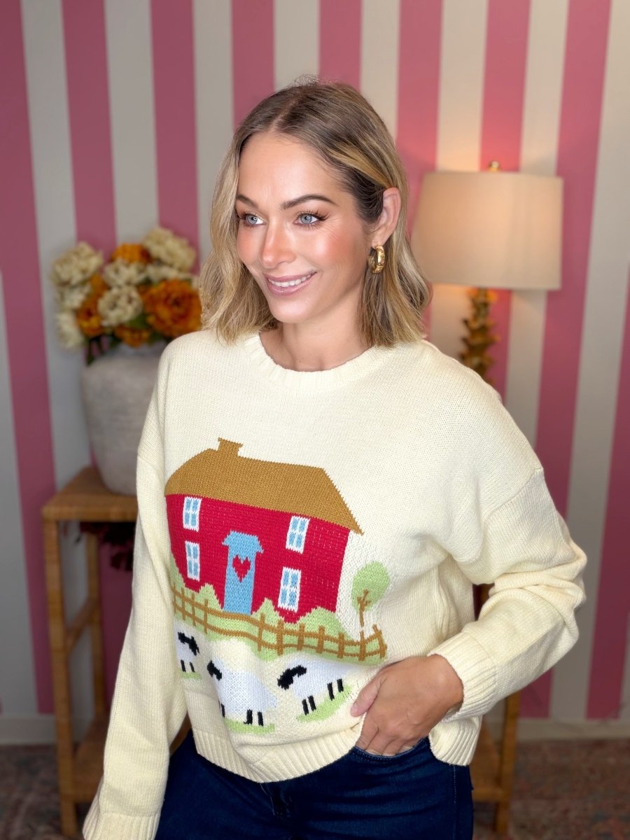 A woman wearing a cream-colored knit sweater with a colorful farmhouse and sheep graphic, posed in front of a pink and white striped wall.