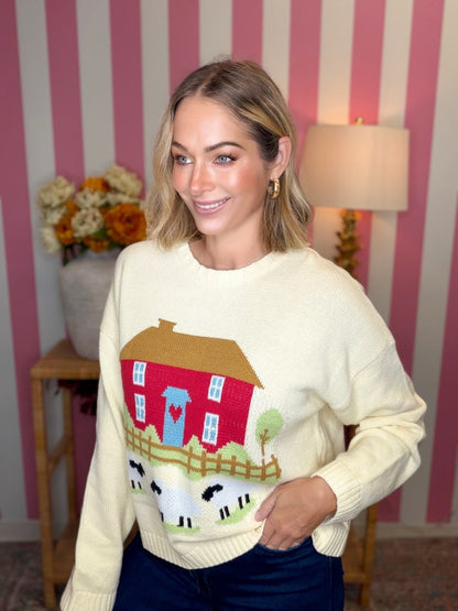 A woman wearing a cream-colored knit sweater with a colorful farmhouse and sheep graphic, posed in front of a pink and white striped wall.