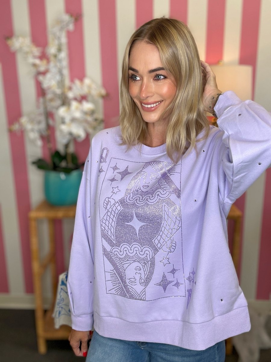 A woman wearing a lavender crewneck sweatshirt with a silver graphic print of a hot air balloon and stars, posed in front of a pink and white striped wall.