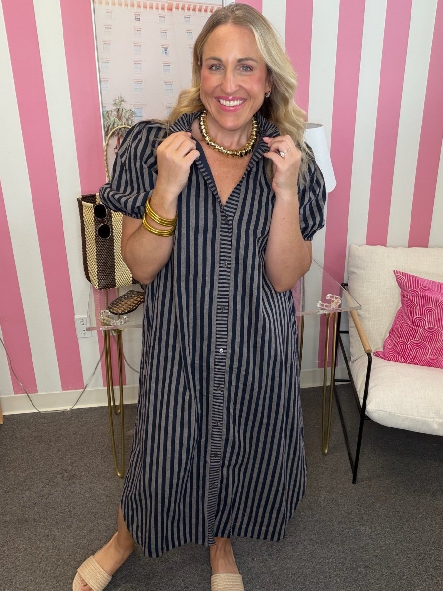 A woman modeling a navy and white vertically striped midi shirt dress with puff sleeves and a button-down front, styled with gold jewelry in a pink and white striped room.