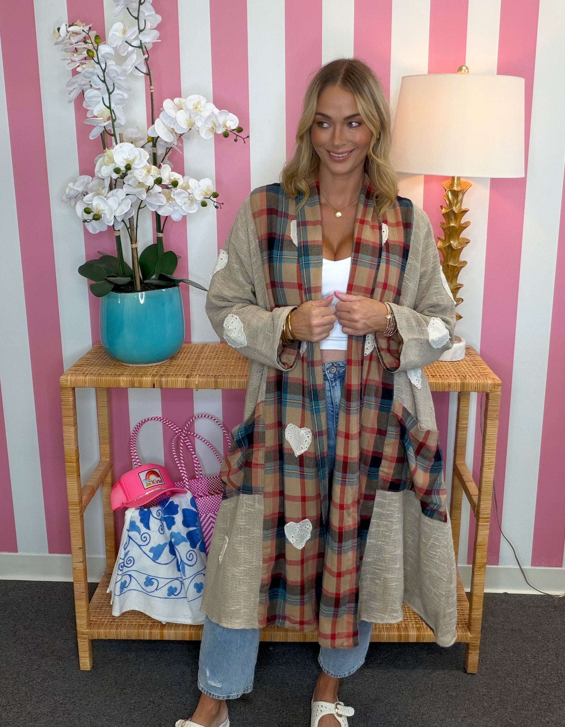 A woman stands in front of a pink and white striped wall wearing a long beige cardigan with a red, blue, and green plaid patchwork front and white heart appliqués. She is standing next to a wooden side table with a turquoise orchid and a lamp.