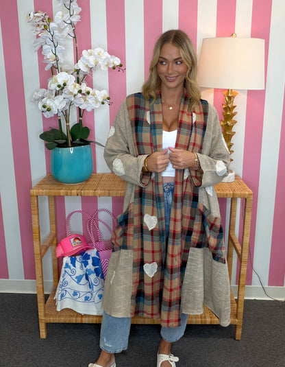 A woman stands in front of a pink and white striped wall wearing a long beige cardigan with a red, blue, and green plaid patchwork front and white heart appliqués. She is standing next to a wooden side table with a turquoise orchid and a lamp.