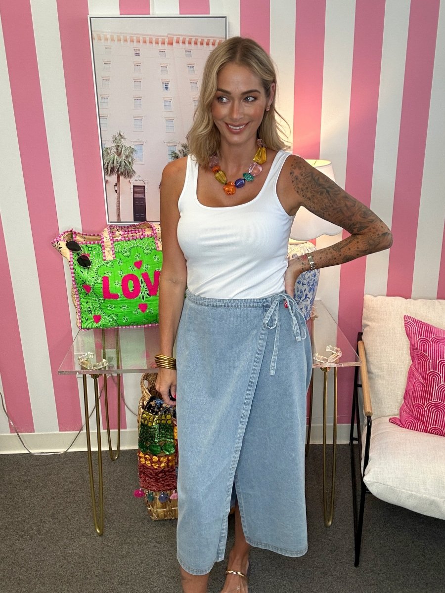 A woman modeling a white ribbed tank top and a light blue denim wrap midi skirt, standing in front of a pink and white striped wall. She is accessorized with a chunky multi-colored beaded necklace and gold bracelets.