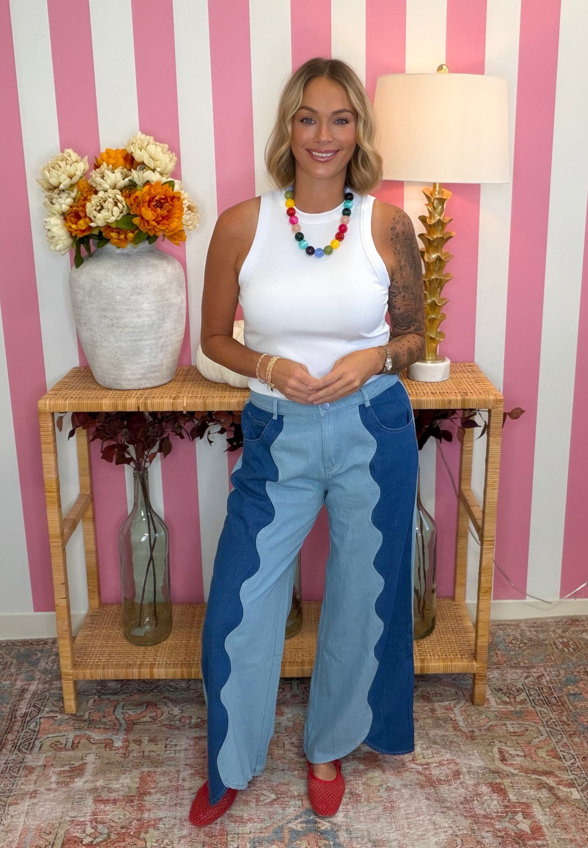 A woman stands in front of a pink and white striped wall wearing a white tank top and distinctive light blue wide-leg jeans with bold wavy darker blue side panels. She is accessorized with a colorful beaded necklace and red shoes, standing behind a wooden console table decorated with a vase of flowers and a lamp.