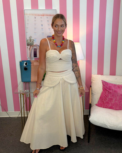 A woman modeling a two-piece cream or off-white knit set consisting of a cropped camisole top and a matching flared skirt, standing in front of a pink and white striped wall.