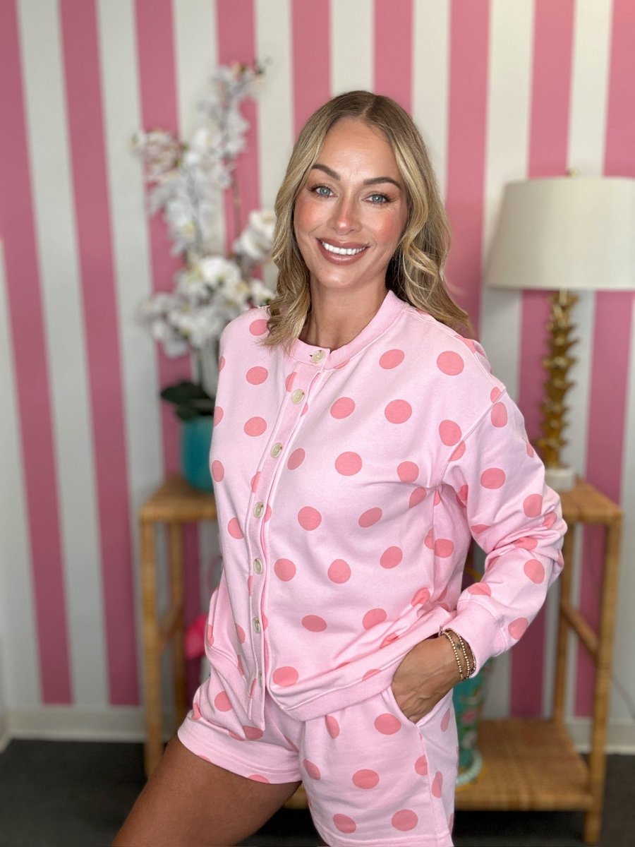 A woman modeling a pink two-piece pajama set with a pink polka dot pattern, consisting of a long-sleeve zip-up top and matching shorts, posed in front of a pink and white striped wall.