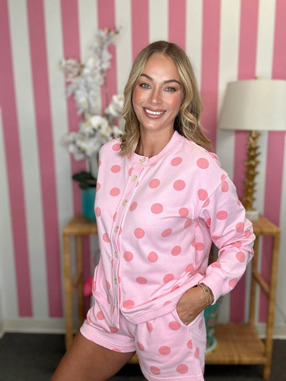 A woman modeling a pink two-piece pajama set with a pink polka dot pattern, consisting of a long-sleeve zip-up top and matching shorts, posed in front of a pink and white striped wall.