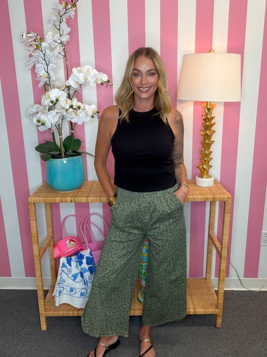 A woman stands in front of a pink and white striped wall, wearing a black sleeveless top and green patterned wide-leg pants. She is posed next to a wooden side table with a turquoise orchid pot and a gold lamp.